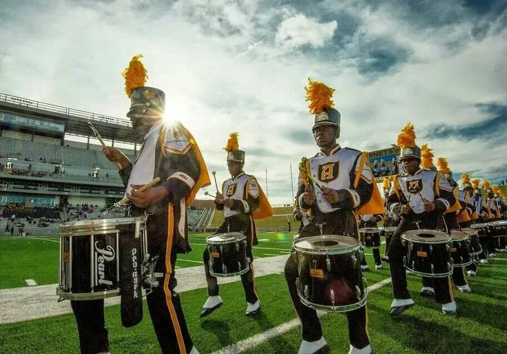 The drumline marches across the field under a bright sky